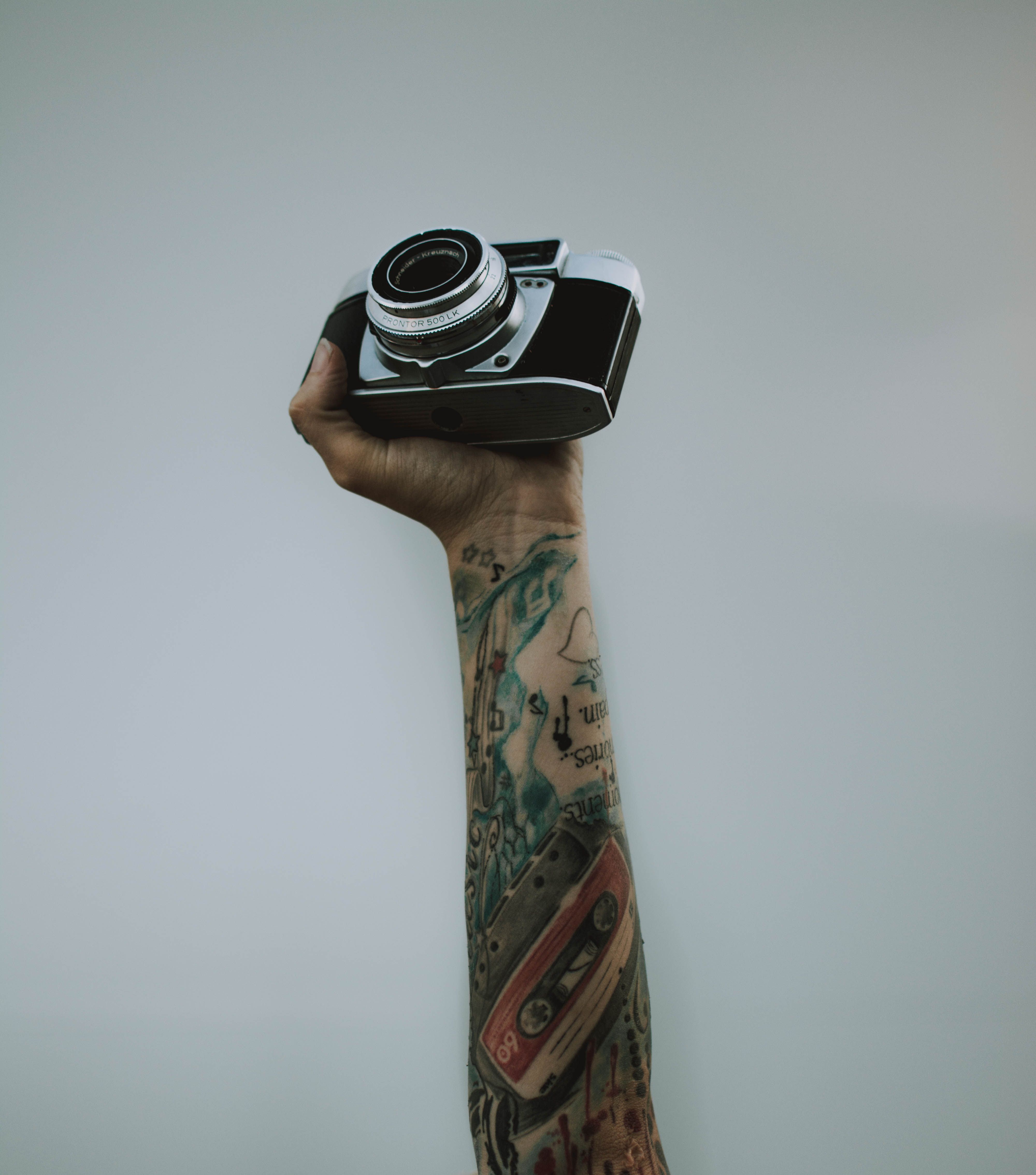 A tattooed arm holding a vintage camera against a plain wall